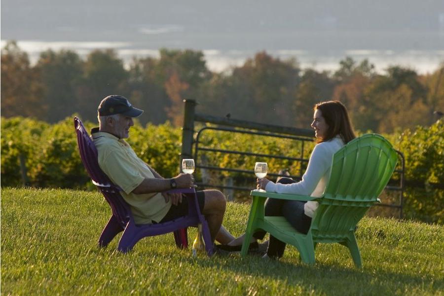 A man and woman sit in Adirondack chairs holding a glass of wine along the hedge of a vineyard.